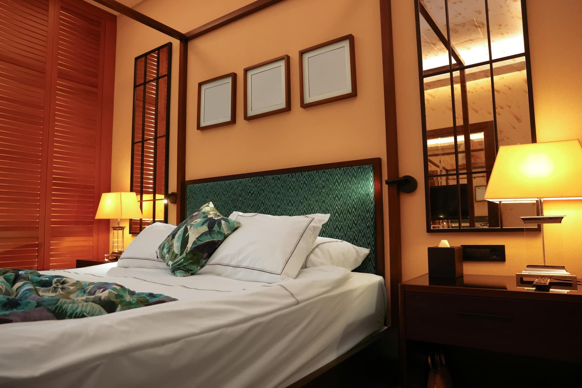 A bed with white pillows and a green patterned headboard, framed by lamps, mirrors and warm orange walls